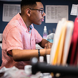 A black man wearing glasses and a short-sleeved pink shirt sits at a desk, focused on his computer screen. The desk is cluttered with various papers and folders, with a row of colorful file folders prominently visible in the foreground.