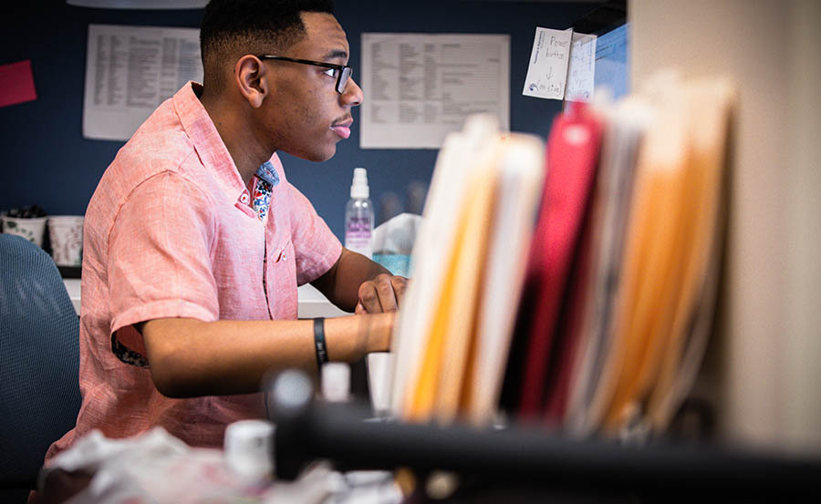 A black man wearing glasses and a short-sleeved pink shirt sits at a desk, focused on his computer screen. The desk is cluttered with various papers and folders, with a row of colorful file folders prominently visible in the foreground.