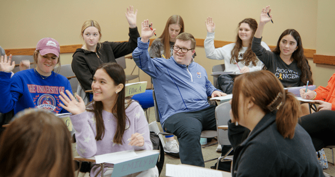 Photo of students in a college classroom, raising hands and smiling, including one male with Down syndrome in the center.