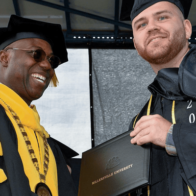 A Black man with a big smile wearing black and gold robes confers a degree to a white male student. The student is facing the audience smiling, holding his diploma. 