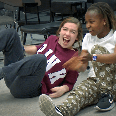 Two girls laughing together on the floor in dance class. They are dressed casually, in sweats, one is leaning back on her arm with her feet off the ground, the other has one leg straight in front of her and the other bent with her arm around it.