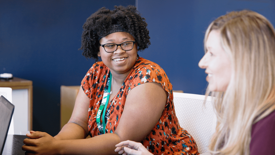 Black female college student smiling and looking to her left at a white female student who is looking across the table at someone else.